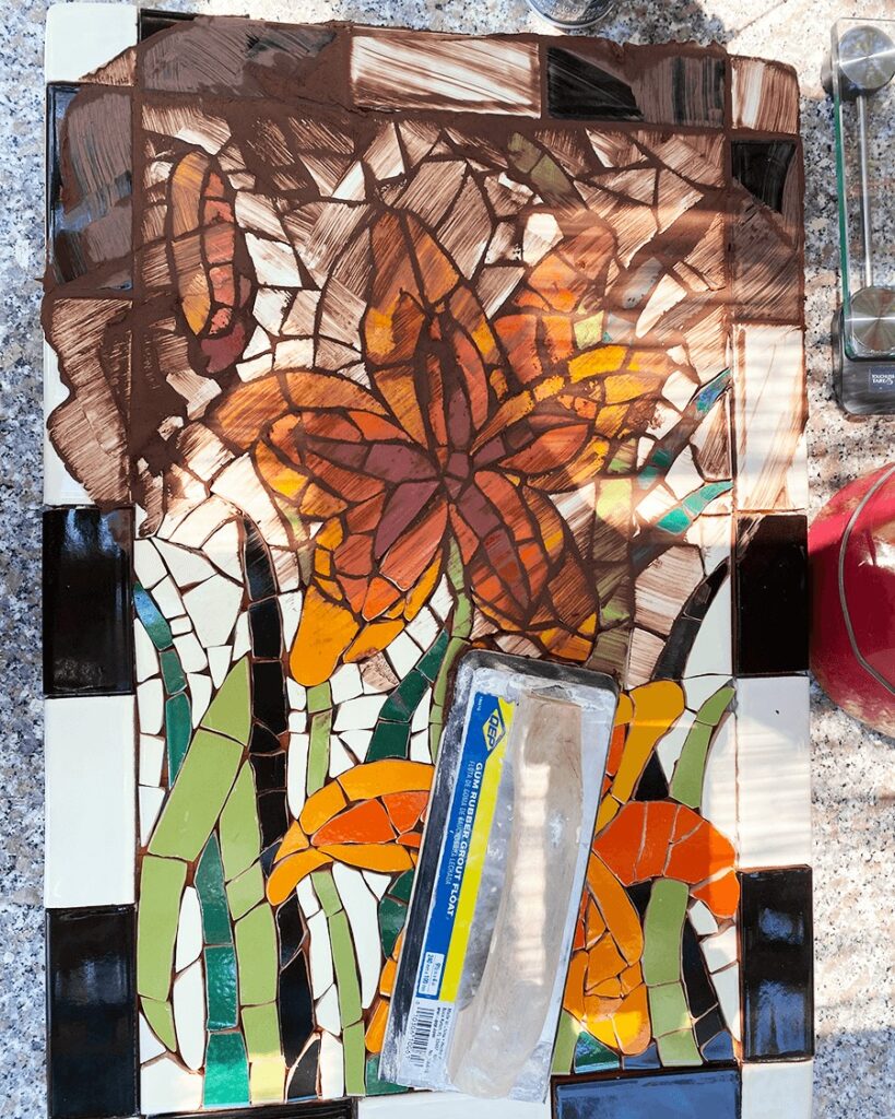 mosaic tabletop half grouted with dark brown grout along side a grout float