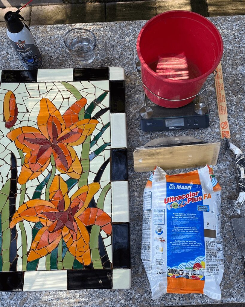 trencadis broken mosaic tile tabletop prepared to grout alongside a bag of mapei grout, a grout float and a bucket